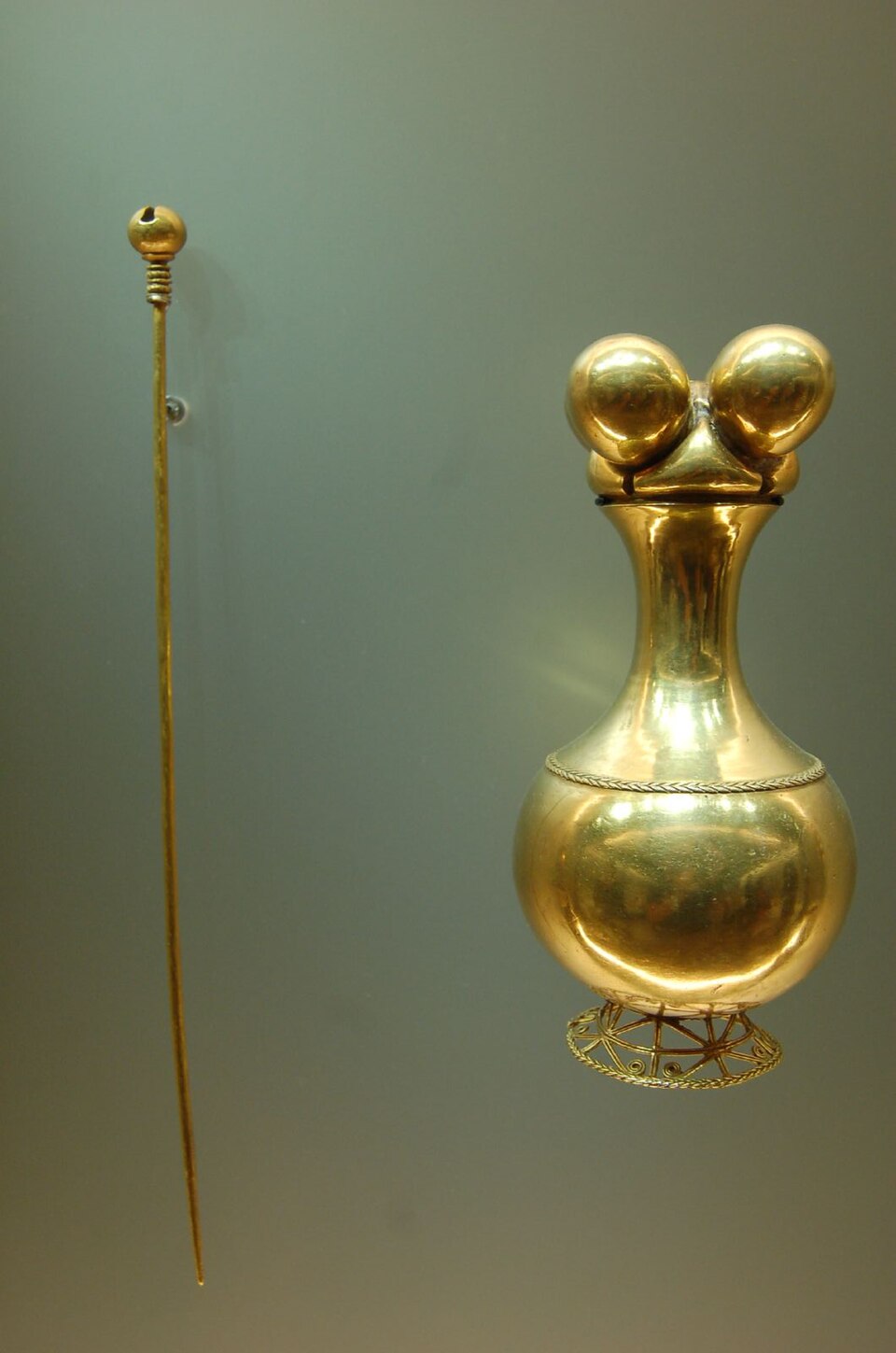 Gold Quimbaya Poporo with a rounded body, slender neck and four spherical lobes, displayed beside a long pin.