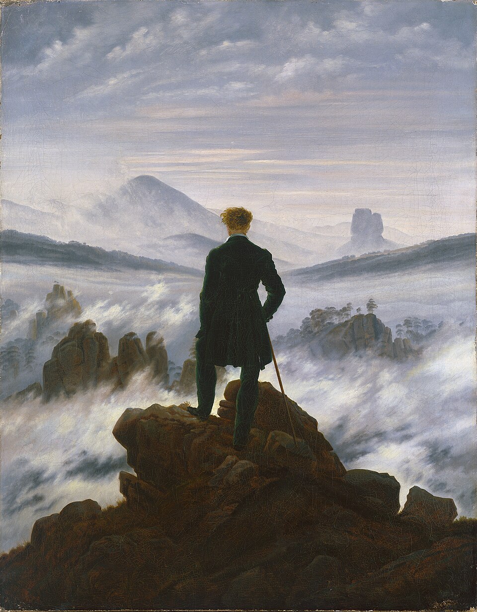 A man viewed from behind stands on a rocky peak above fog-covered mountains.