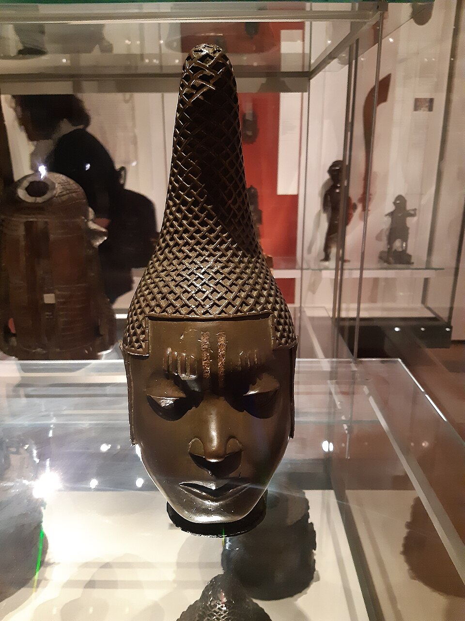 Bronze commemorative head of Queen Idia with a tall lattice headdress, displayed in a museum case.