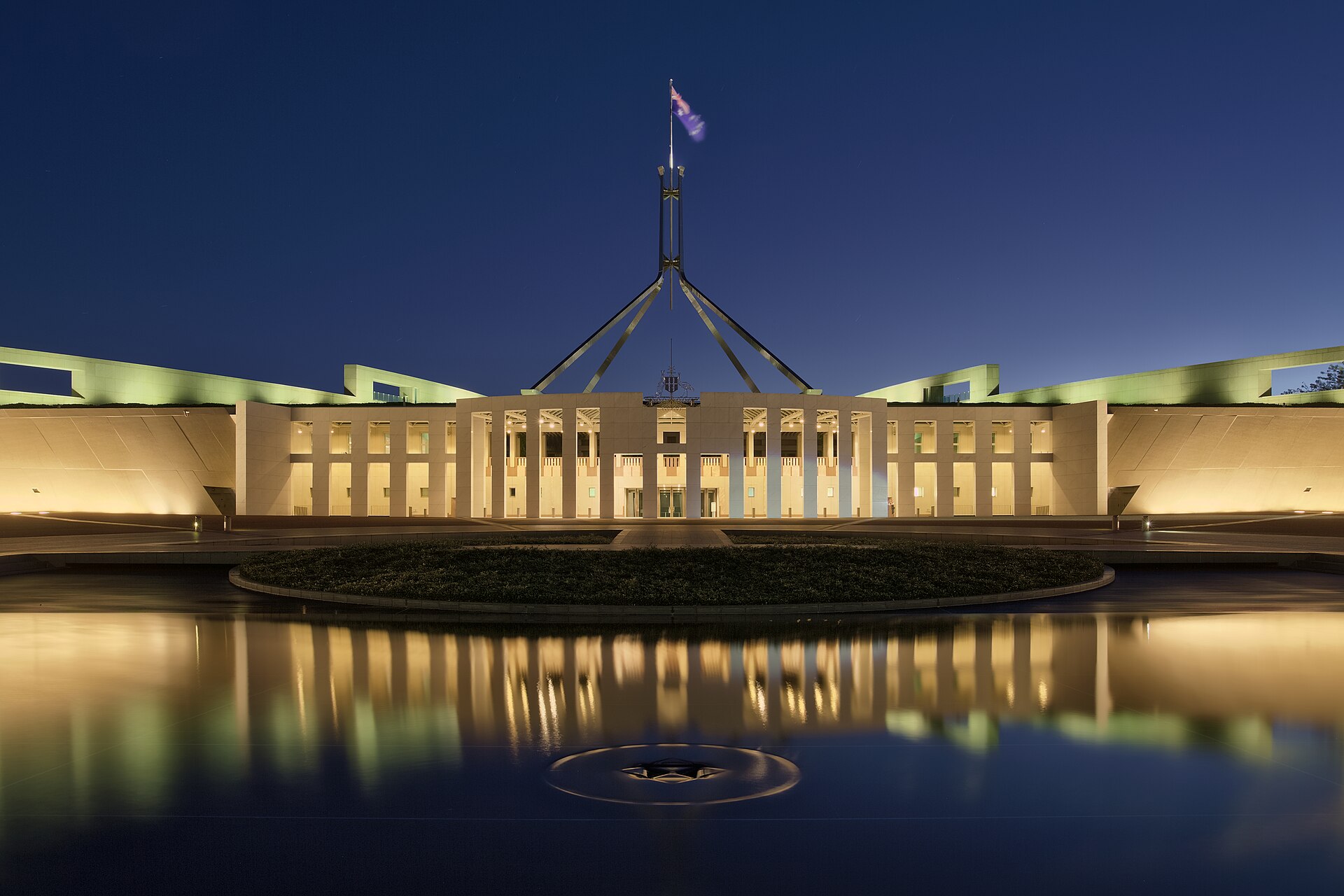 Illuminated Australian Parliament House at dusk, reflected in water.