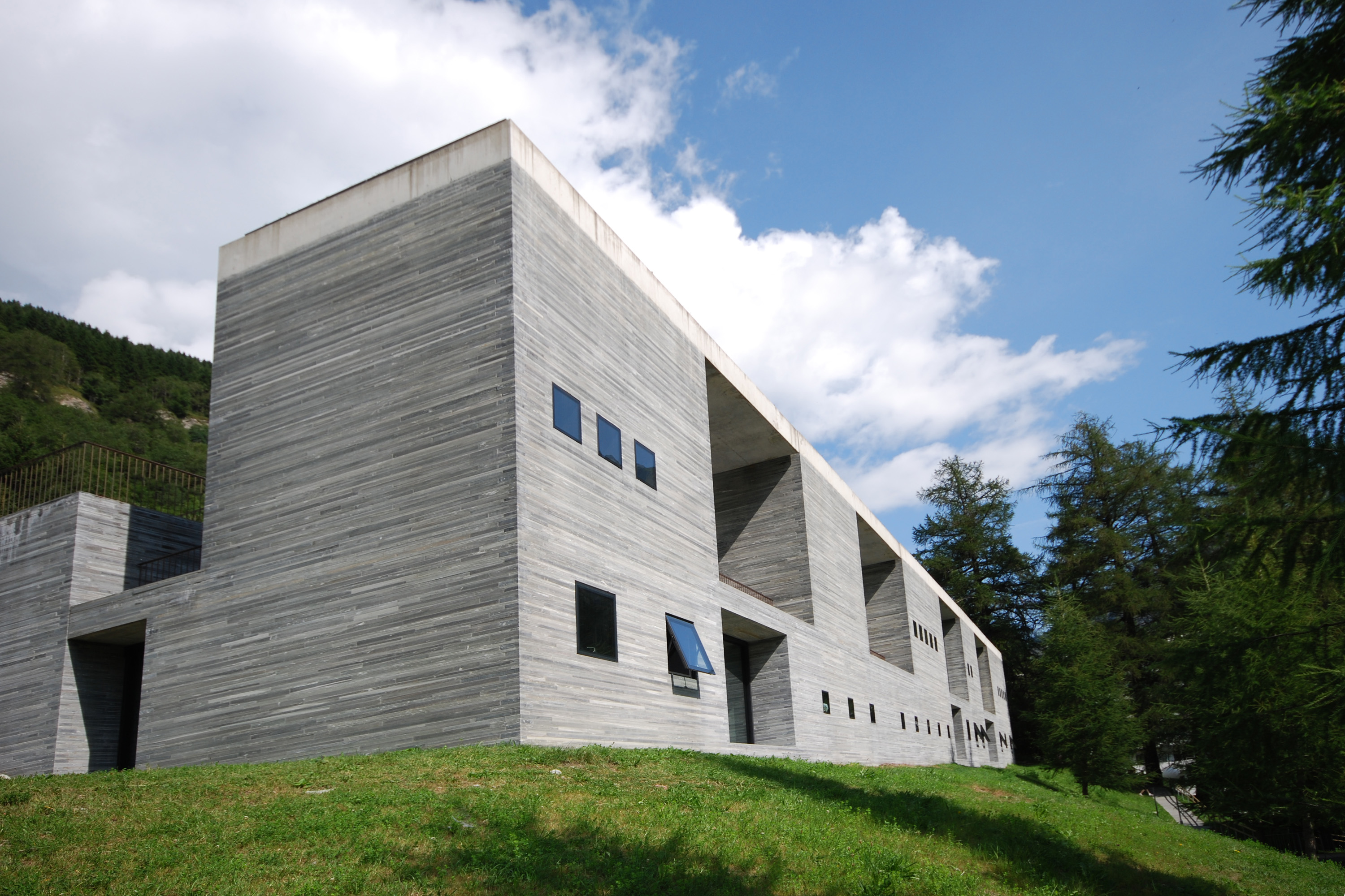 Rectangular stone facade of Therme Vals with small windows beside a grassy slope.