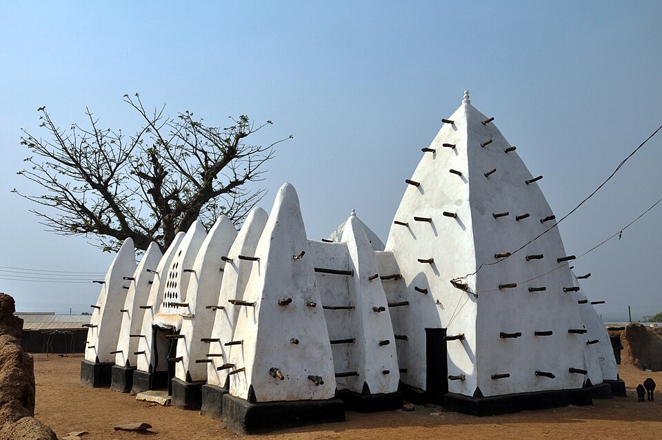 White earthen mosque with projecting timber elements and a tree beside it.