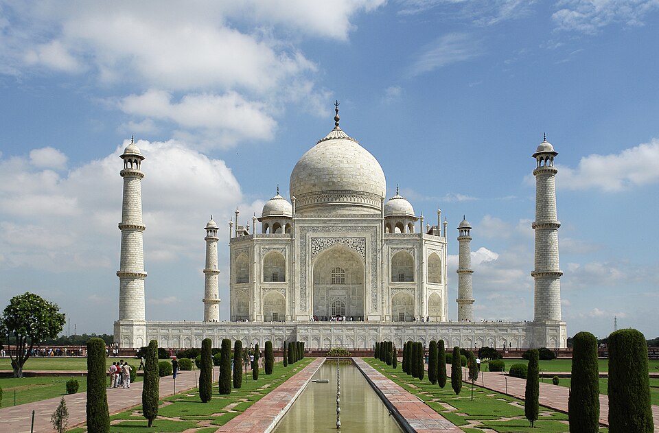 The white marble Taj Mahal and four minarets, seen along the central garden and reflecting pool beneath a blue, clouded sky.