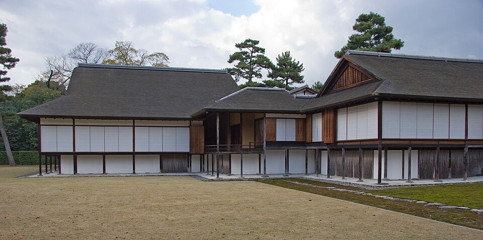 Low Japanese timber buildings with sliding screens facing a flat grassy courtyard.