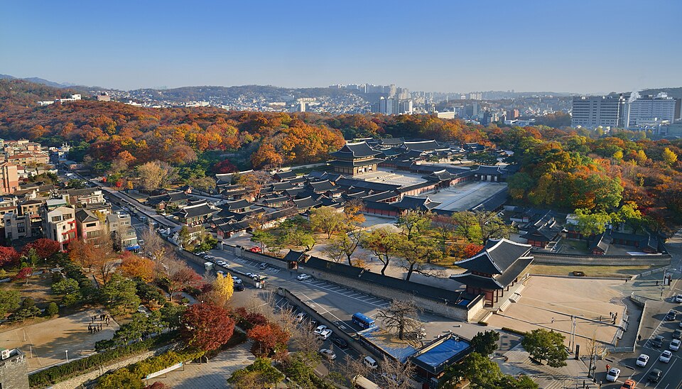Palace roofs and courtyards surrounded by autumn trees, with a city skyline beyond.