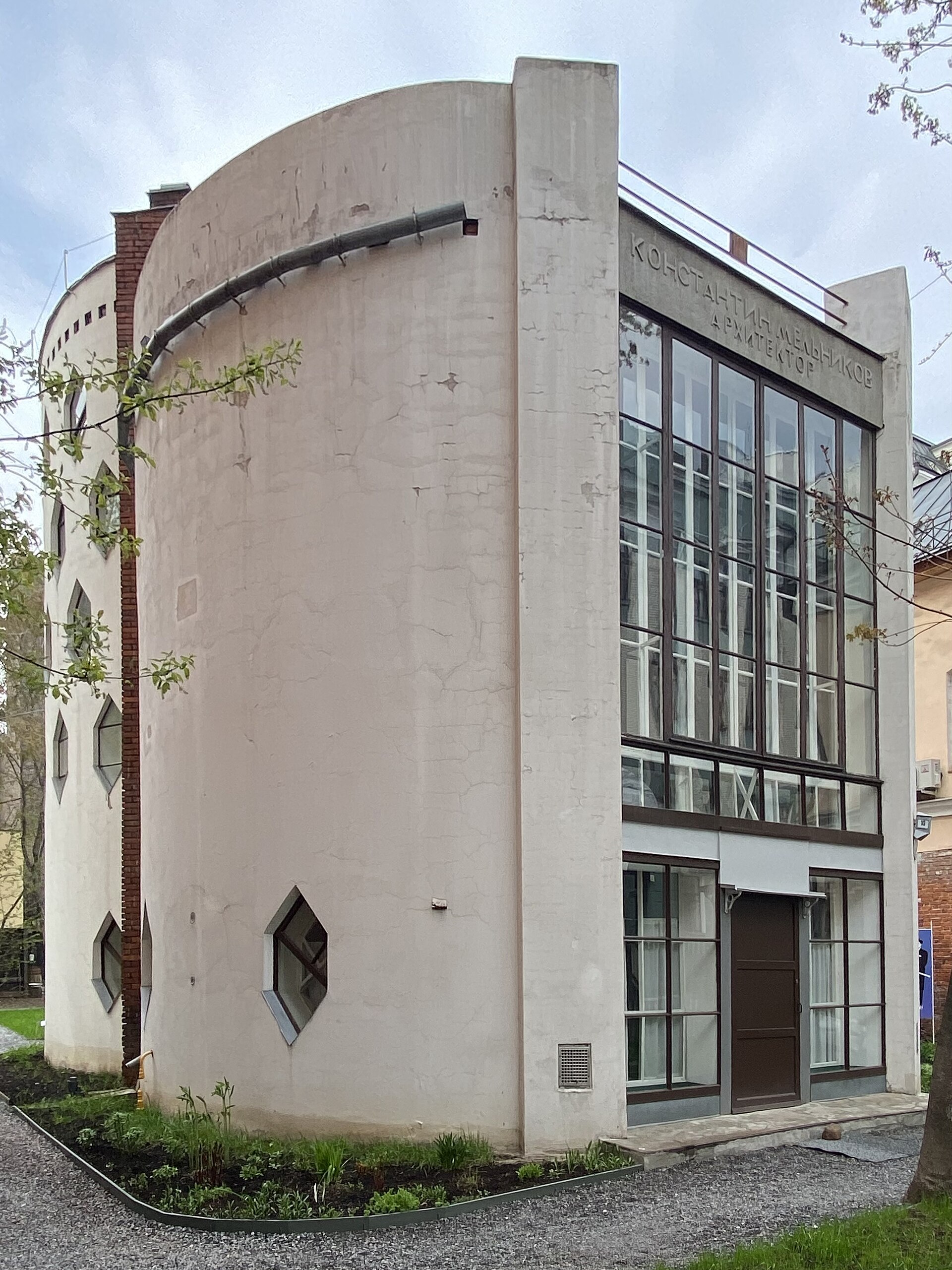 Cylindrical Melnikov House with a tall glazed facade, partly screened by trees.